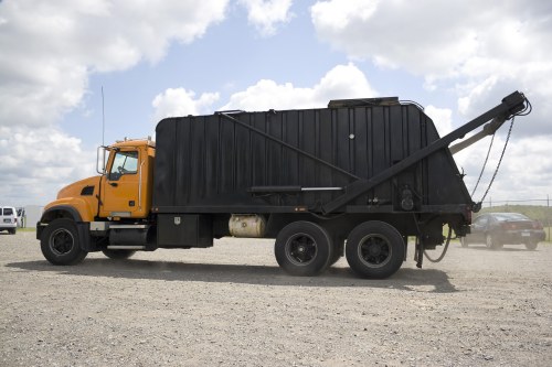 A large commercial waste collection truck with an orange cab and black waste container, parked on a gravel surface under a partly cloudy sky. The container appears to be made of metal with a ribbed texture and is fitted with an hydraulic lifting mechanism at the rear, used for tipping or unloading rubbish. The truck features six wheels, with two at the front and four at the rear, and is positioned in an outdoor setting that suggests a waste disposal or recycling site near Great Bookham. In the background, additional parked vehicles are visible on the right side. The scene is well-lit by daylight, highlighting the industrial design of the vehicle, which is used by companies like Commercial Waste Great Bookham for efficient rubbish removal services in the local area.