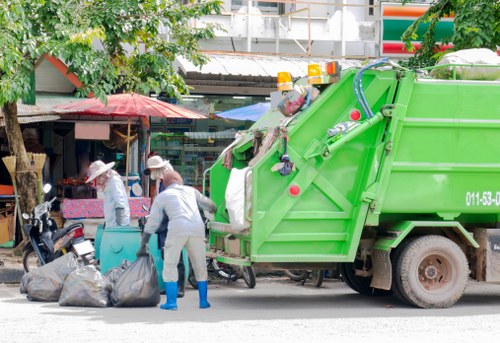 A green commercial rubbish collection vehicle parked on the side of a street in a residential or urban area, with workers dressed in light grey uniforms and protective gloves lifting black garbage bags onto the vehicle's rear loading area. The vehicle has a hydraulic lifting mechanism visible on the side. In the background, there are small shops or market stalls with colorful umbrellas or awnings, and a few pedestrians, some of whom wear hats. The scene occurs in daylight, with trees and building structures visible behind the vehicle, indicating a typical rubbish removal operation in Great Bookham or nearby areas in Surrey.