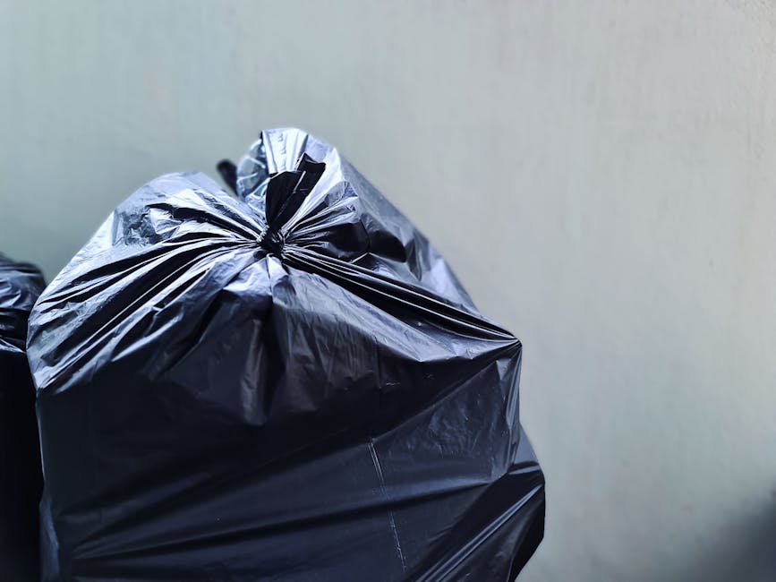 Close-up image of several black plastic rubbish bags filled with waste, tied securely at the top, and placed against a plain, light-colored wall. The bags appear to be made of shiny, crinkled plastic