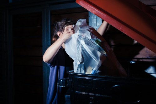 A person wearing a dark shirt is seen disposing of clear plastic rubbish bags filled with waste into a large black skip or container, which is situated outdoors on a paved area adjacent to a wooden structure. The scene is dimly lit, emphasizing shadows and focusing light on the individual and the waste materials. The waste bags appear to contain mixed rubbish, possibly including paper or plastic items, and are tightly sealed at the top. The black skip is positioned close to a red-colored door or lid, which the person is holding open with their right hand while depositing the bags. The background features dark blue or black metal framework and wooden paneling, suggesting an environment typical of commercial or industrial waste collection sites consistent with rubbish removal services in Great Bookham, as provided by Commercial Waste Great Bookham.